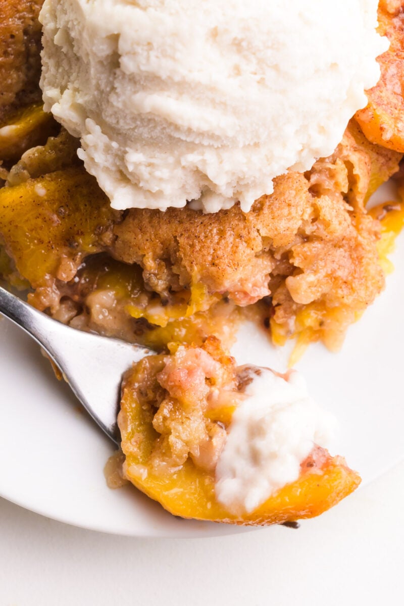 A forkful of vegan peach cobbler sits in front of the rest of the cobbler on a plate. It's topped with vegan vanilla ice cream.