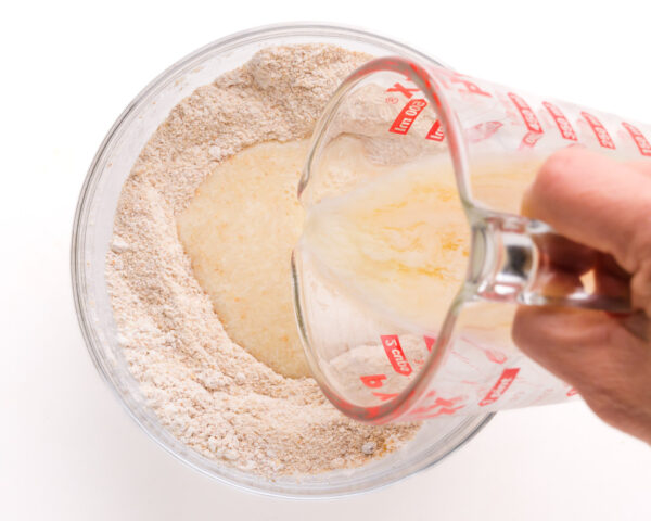 A hand holds a pyrex measuring cup, pouring liquid into a bowl with flour.