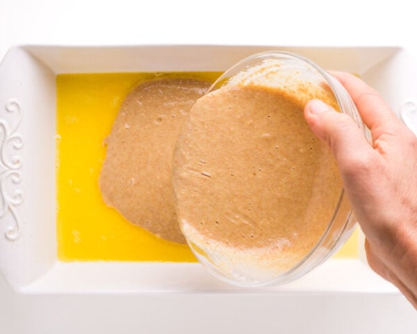 A hand holds a bowl pouring batter into a baking dish with melted butter.