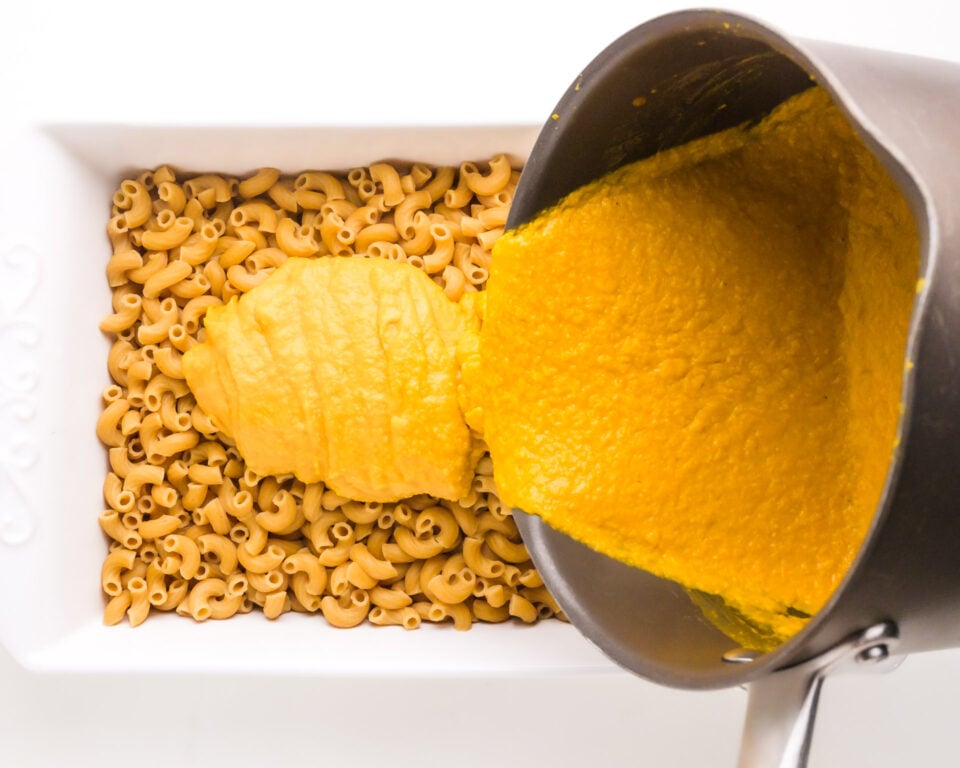 Chickpea cheese sauce is being poured from a saucepan to a casserole dish with cooked macaroni.