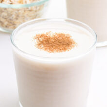 A glass of oat milk sits in front of another glass and a bowl of oats.