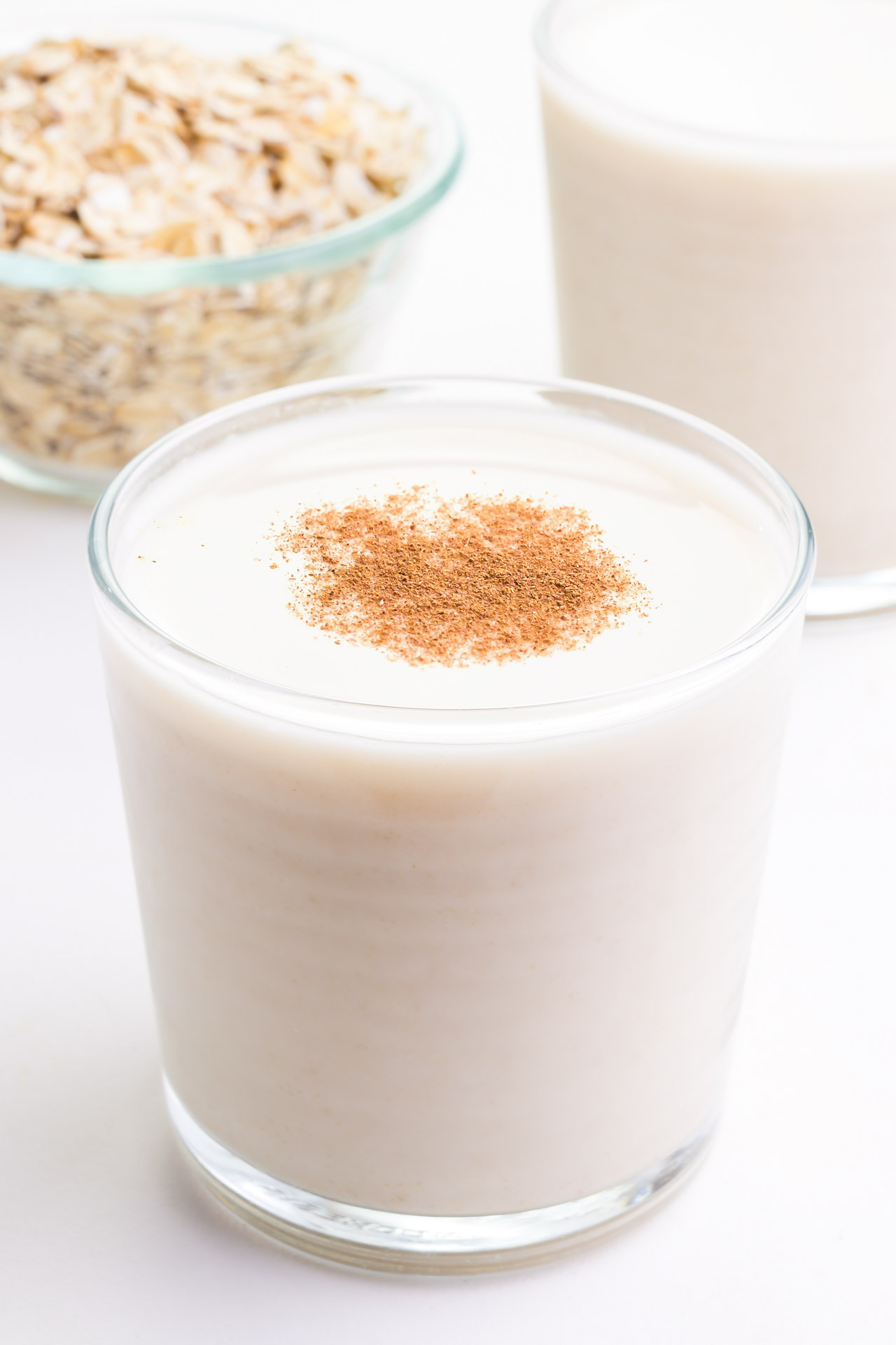 A glass of oat milk sits in front of another glass and a bowl of oats.