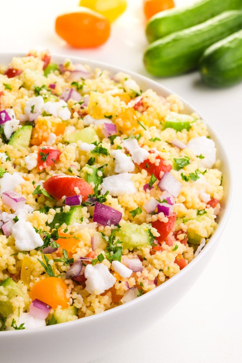 A bowl is full of vegan couscous salad. There are cucumbers and cherry tomatoes in the background.