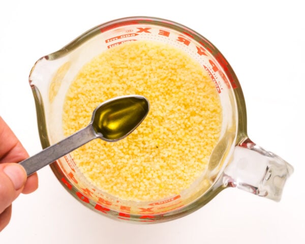 A hand holds a spoonful of olive oil over couscous in a measuring cup.