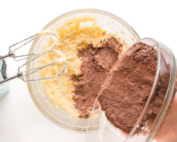 Chocolate flour mixture is being poured into a mixing bowl with whipped butter ingredients.