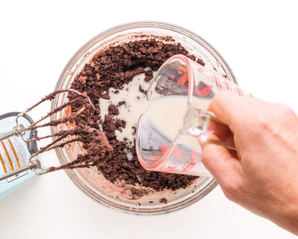 Milk is being poured into a bowl with chocolate cookie batter. There is a mixer sitting by the bowl.