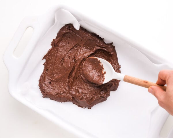 A spatula spreads chocolate batter into a white baking pan.