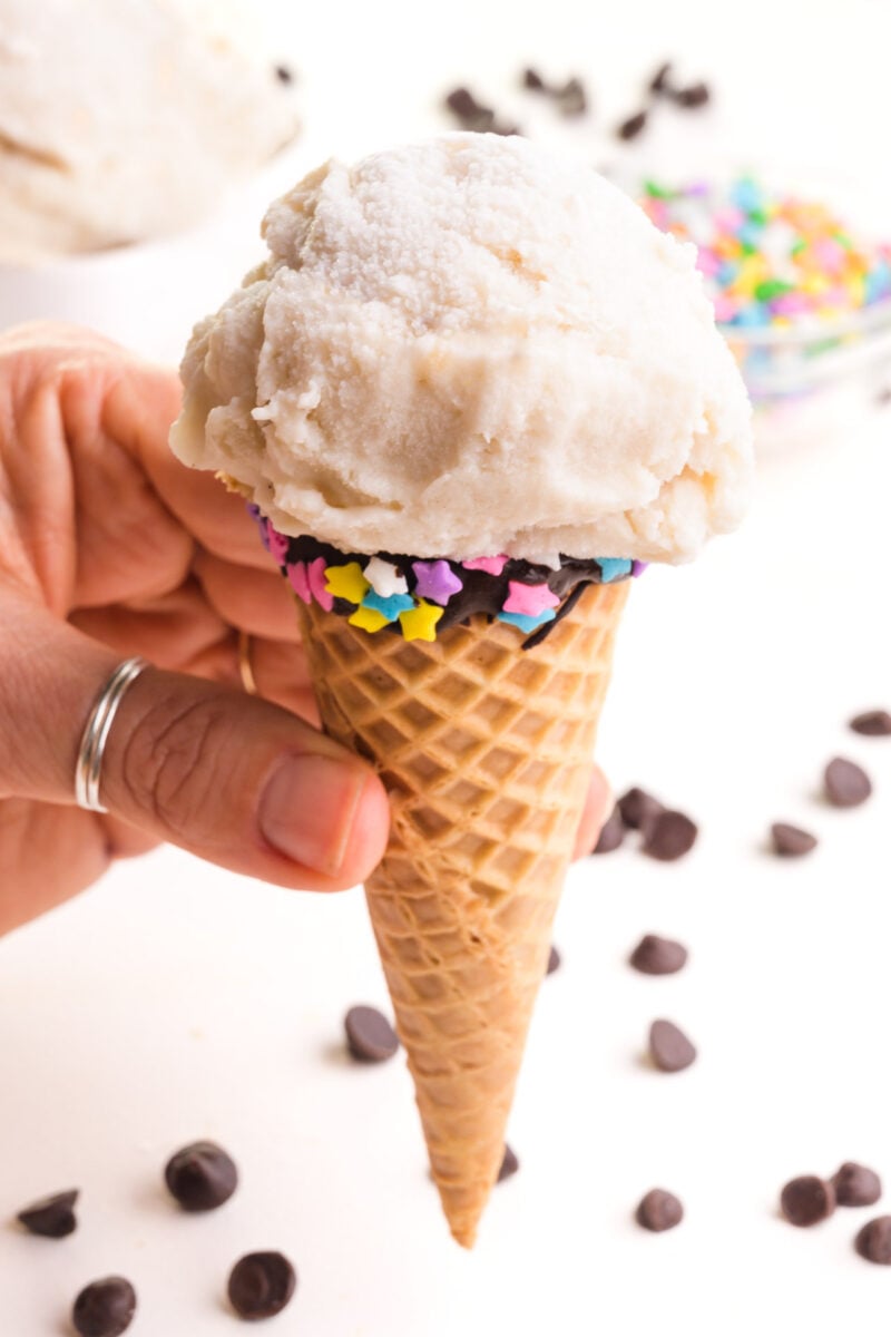 A hand holds an ice cream cone with a scoop of vegan vanilla ice cream.