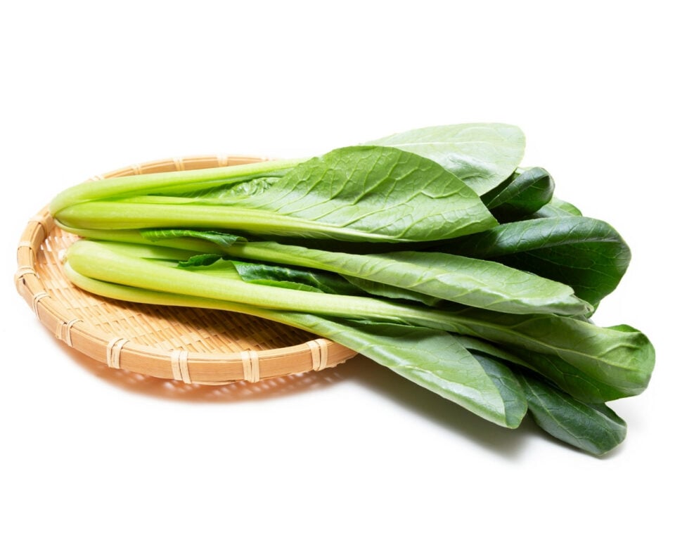 Several stalks of green komatsuna are sitting on a wicker basket on a white counter.