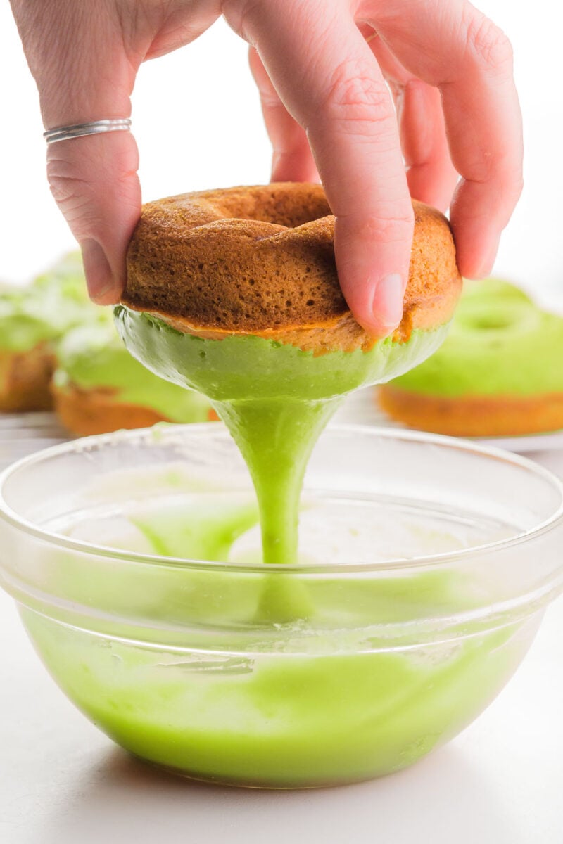A hand holds a donut, dipping it in green frosting. There are more green frosted donuts in the background.