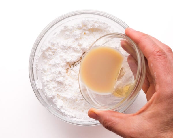 A hand pours liquid from a small bowl into a larger bowl with powdered sugar.