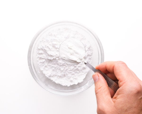 A hand holds a spoonful of cornstarch, getting read to pour it into a bowl with powdered sugar.
