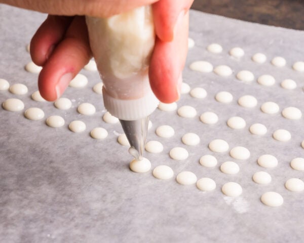 A hand holds a piping bag, piping dots of white icing across a sheet of waxed paper on a pan.