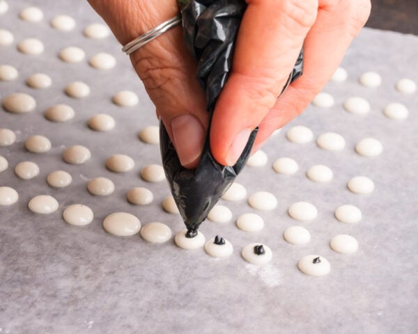 A hand holds a ziplock bag with black icing, adding pupils to white icing dogs on a lined baking sheet.