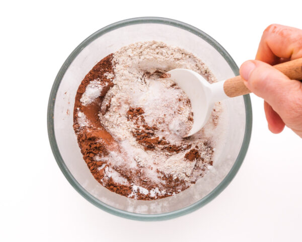 A hand stirs dry ingredients, like flour and cocoa powder, in a mixing bowl.