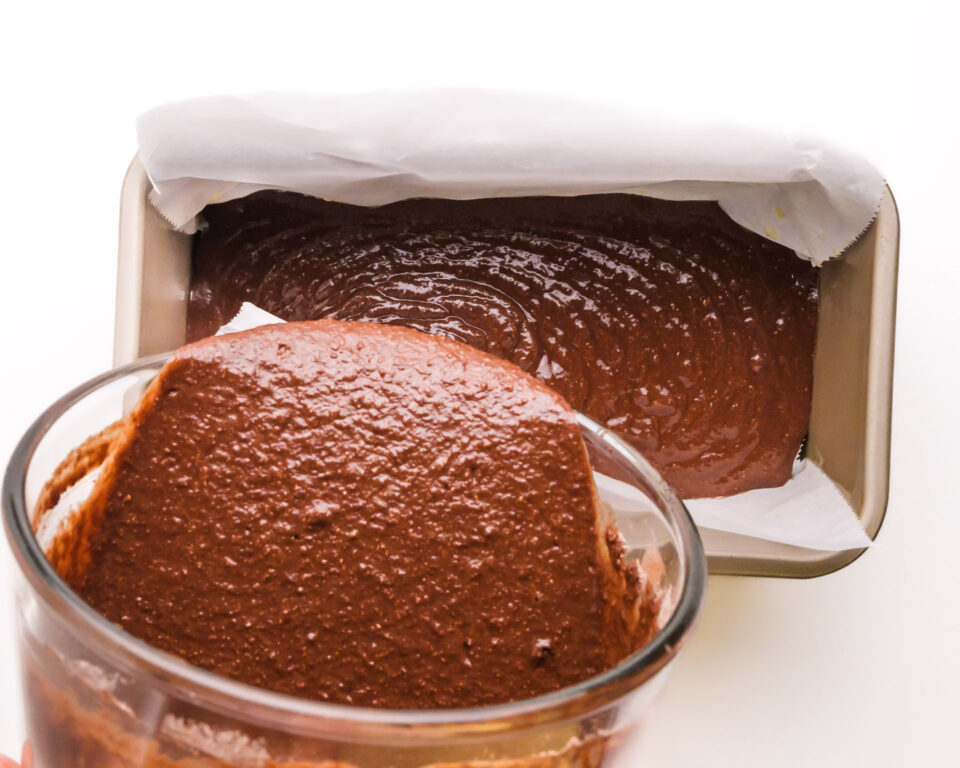 Chocolate batter is being poured from a bowl into a loaf pan.