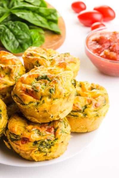 A stack of vegan egg bites on a plate sit in front of greens, cherry tomatoes, and a bowl of salsa.