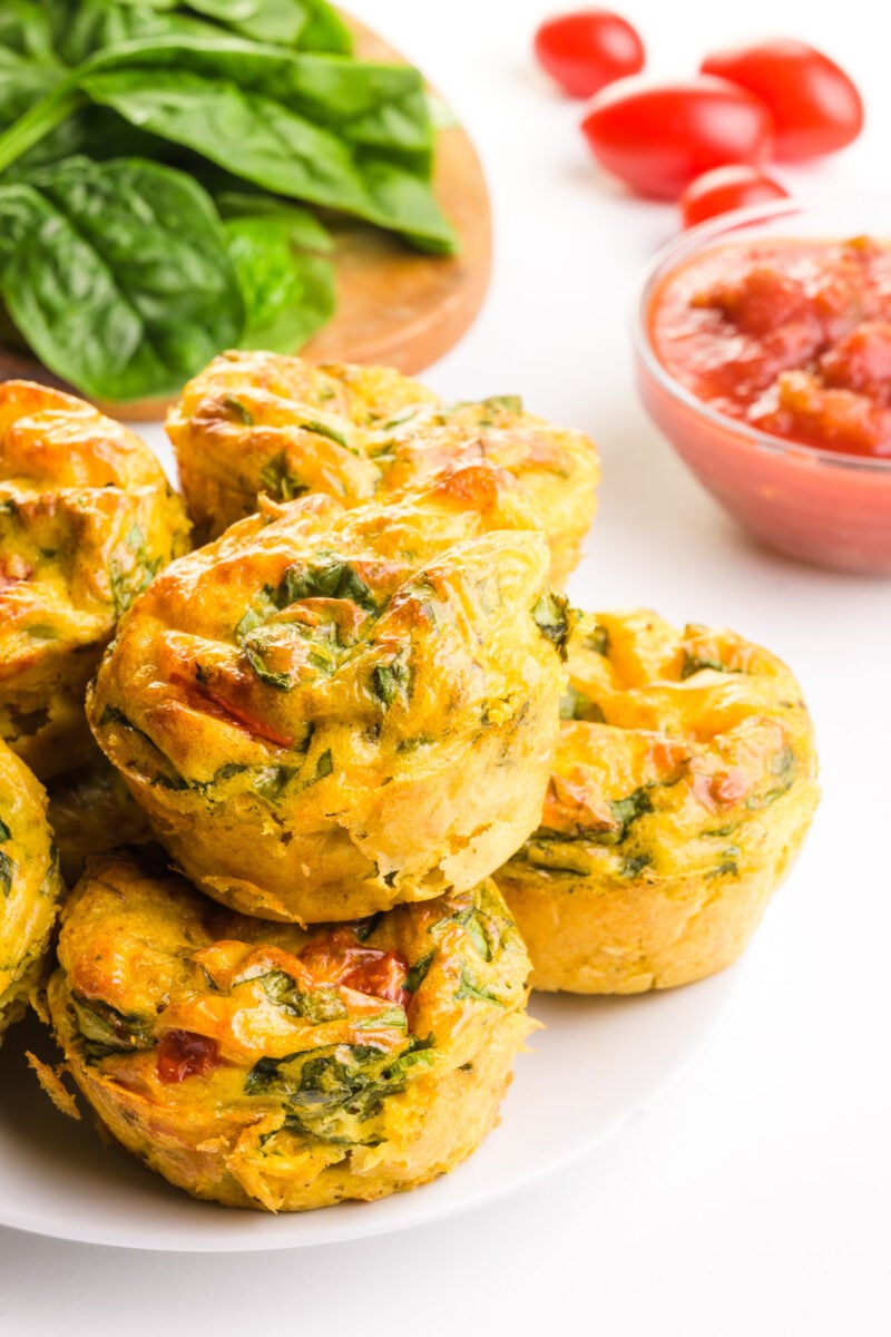 A stack of vegan egg bites on a plate sit in front of greens, cherry tomatoes, and a bowl of salsa.