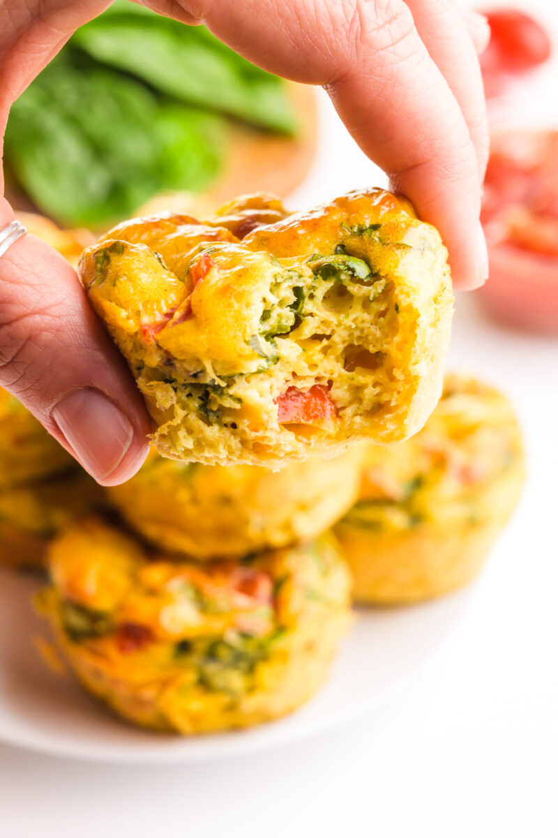 A hand holds a vegan egg bite with a bite taken out. There are more on a plate in the background.