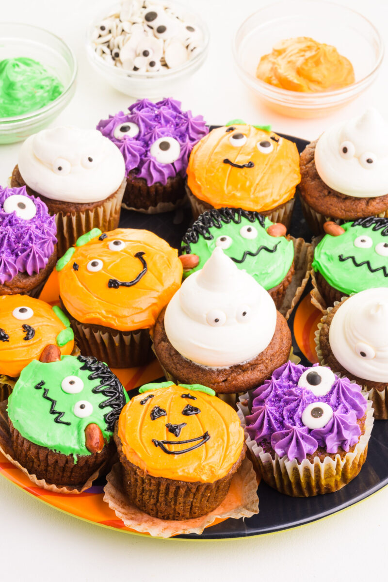 A plateful of colorful vegan halloween cupcakes sits in front of green and orange frosting bowls and googly eyes.