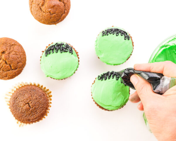 A hand pipes black frosting on green cupcakes. Some plain cupcakes sit beside them.