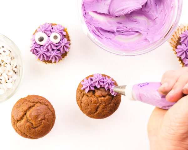 A hand pipes purple frosting stars on cupcakes. One has candy eyes on it and it sits next to a bowl of purple frosting.
