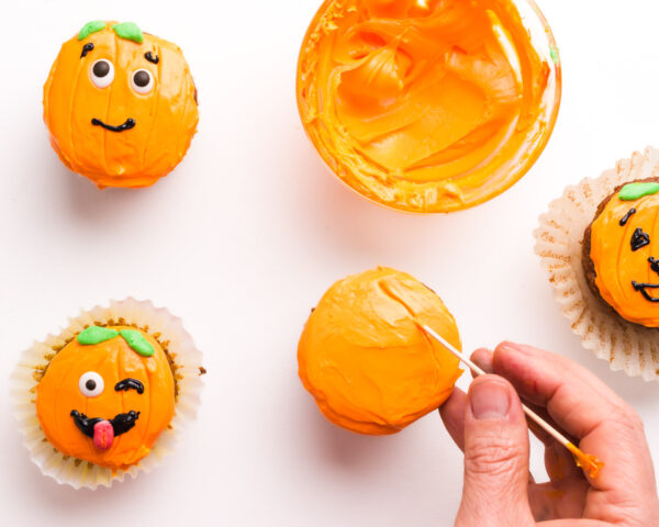A hand uses a toothpick to etch lines in orange frosting on a cupcake to make it look like a pumpkin.