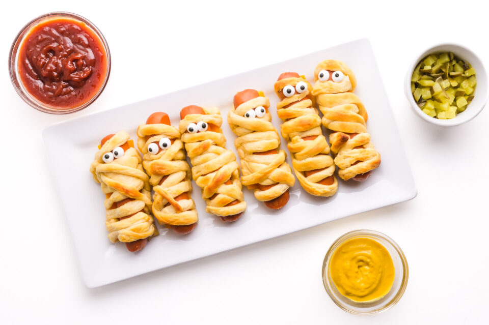 Looking down on a platter of vegan mummy dogs sitting beside bowls of condiments.