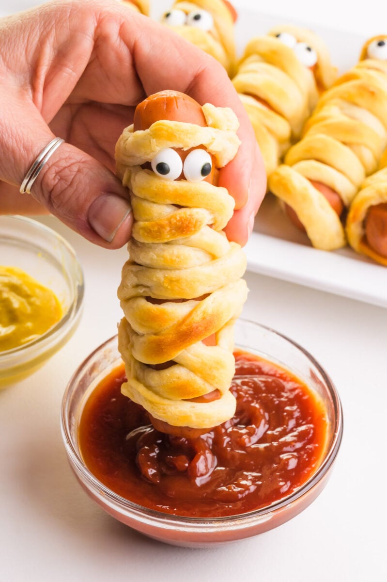 A hand holds a mummy dog with candy eyes, dipping it into a bowl of ketchup. There are more of the dogs in the background.