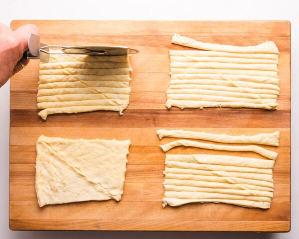A hand uses a pizza cutter to cut rectangles of crescent dough into thin strips.