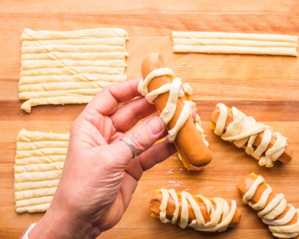 A hand holds a vegan hot dog, wrapping it in crescent roll strips. It hovers over a cutting board with more ingredients.