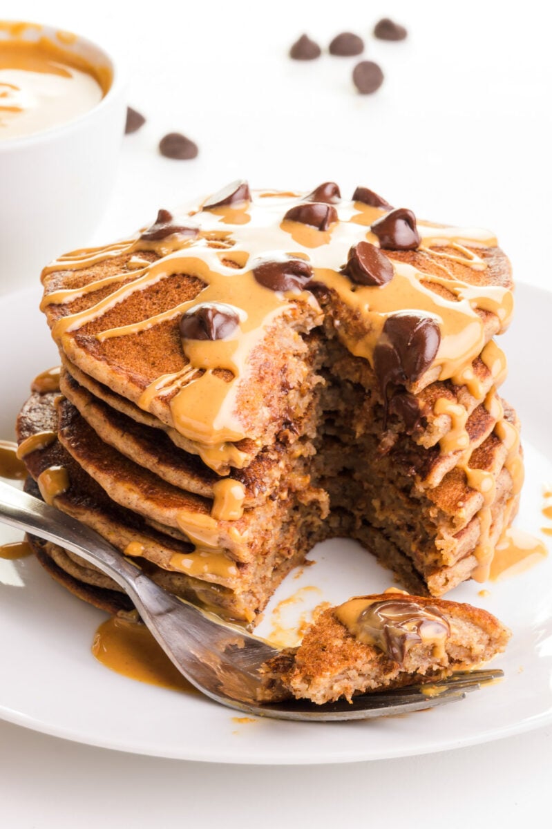 A fork holds a bite of pancake sitting in front of a stack of pancakes with a bite taken out. 