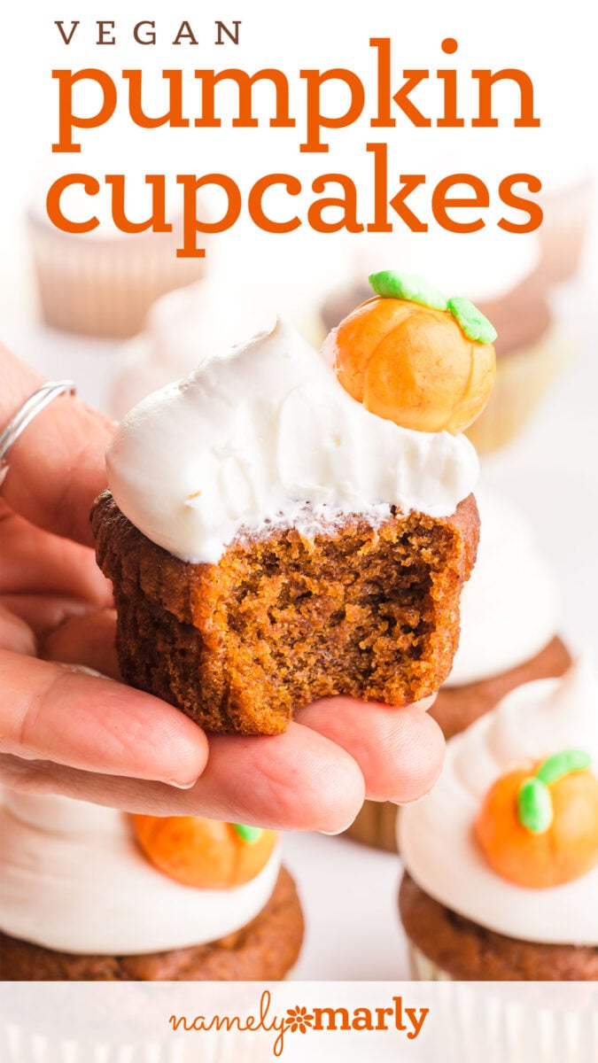 A hand holding a vegan pumpkin cupcake topped with frosting and a decorative mini pumpkin.