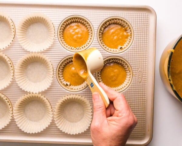 A hand pours pumpkin batter into muffin pan compartments.