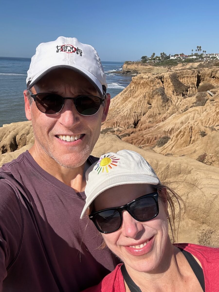 A couple stand in front of a cliff near the Pacific Ocean.