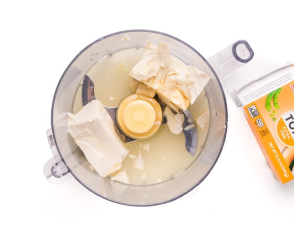 A food processor holds tofu and other ingredients. It sits beside an empty box of silken tofu.