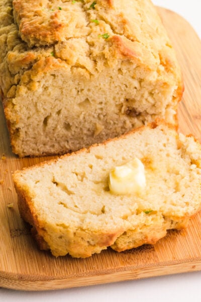 A loaf of vegan beer bread sits on a cutting board. There is a slice in front of it with a pat of butter on it.