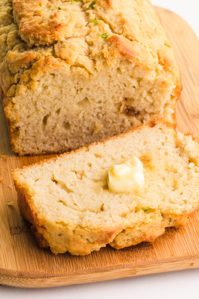 A loaf of vegan beer bread sits on a cutting board. There is a slice in front of it with a pat of butter on it.