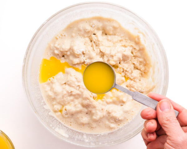 A hand holds a measuring spoon of melted butter, pouring it into a bowl with batter.