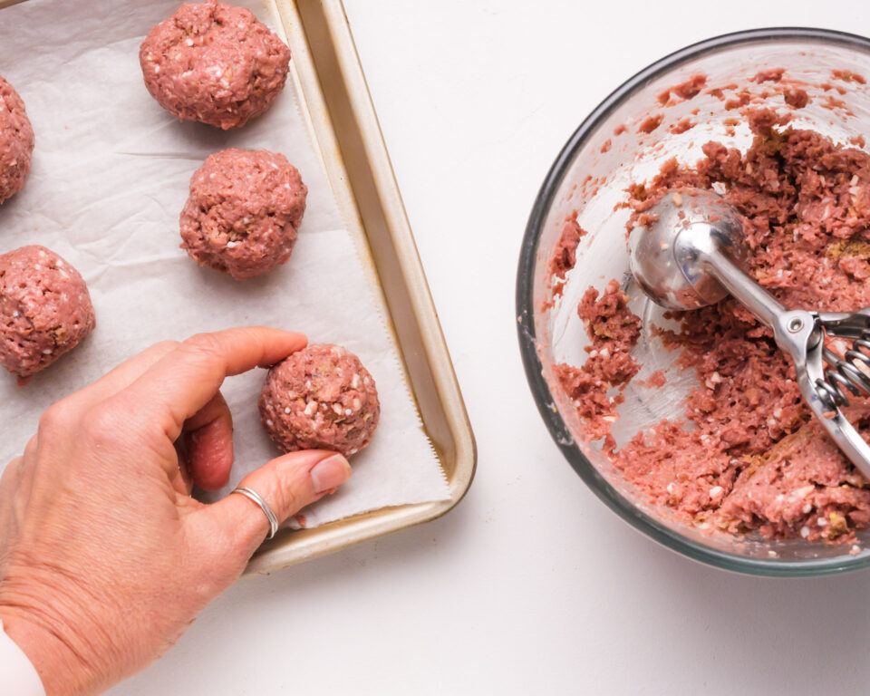 Hands at work in the kitchen as uncooked Impossible Meat Meatballs are carefully placed on a baking sheet. A bowl filled with the flavorful meatball mixture and a convenient dispenser sit nearby, highlighting the easy and enjoyable process of preparing these plant-based delights.