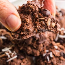 A hand holds a no-bake high protein cookie with a bite taken out. It hovers over a plate with more of the cookies.