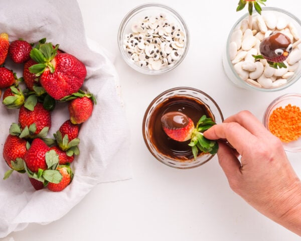 A hand holds a strawberry, dipping it in melted chocolate. There is a plate of strawberries and bowls of candy eyes next to the melted chocolate.