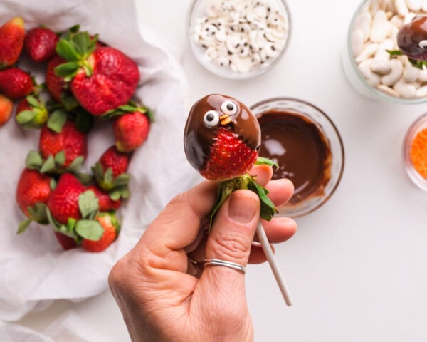 A hand holds a stick with a chocolate dipped strawberry with two candy eyes on it. There is a plate of strawberries and bowls of candies below it.
