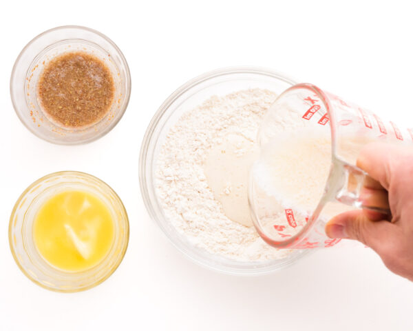 Liquid ingredients are being poured into a mixing bowl with flour. There are small bowls with melted butter and a flax egg beside the mixing bowl.