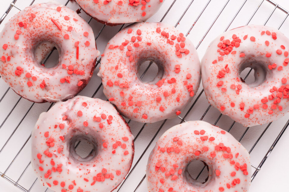 Looking down on several vegan donuts with white glaze and colorful sprinkles on top. They are sitting on a wire rack.