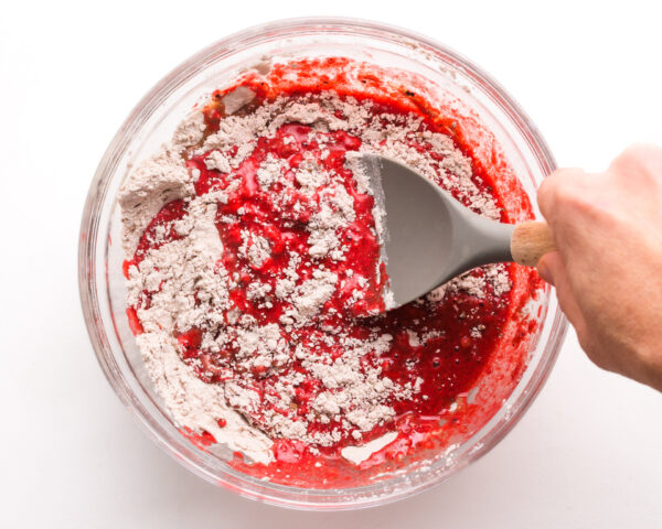 A hand holds a spatula stirring red velvet batter in a mixing bowl.