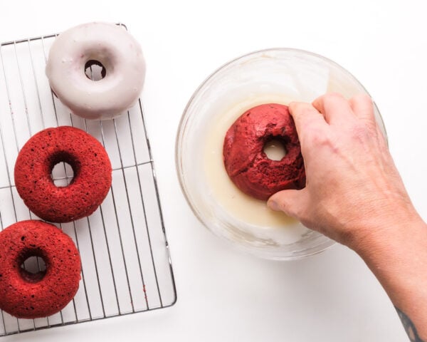 A hand dips a baked red velvet doughnut into icing. There is a wire rack next to it with more donuts.