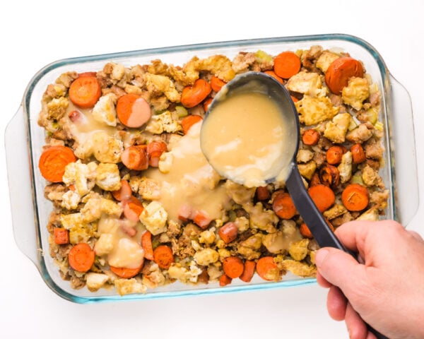 A hand holds a ladle, drizzling gravy over the top of a casserole with carrots and tofu.