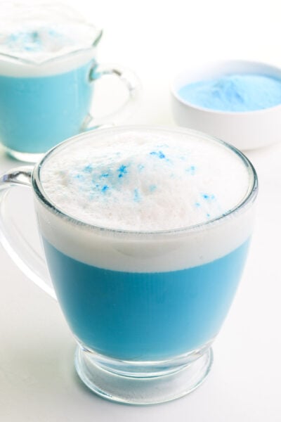 A cup of blue matcha latte sits in front of another glass and a bowl of blue matcha powder.