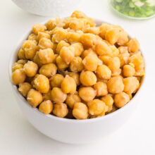 A bowl of cooked chickpeas sits on a counter in front of herbs.